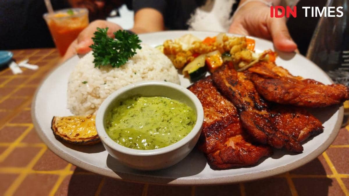 Sepiring hidangan African Brick Chickenette dengan nasi, ayam panggang berbumbu, saus hijau, dan sayuran panggang di atas meja makan.
