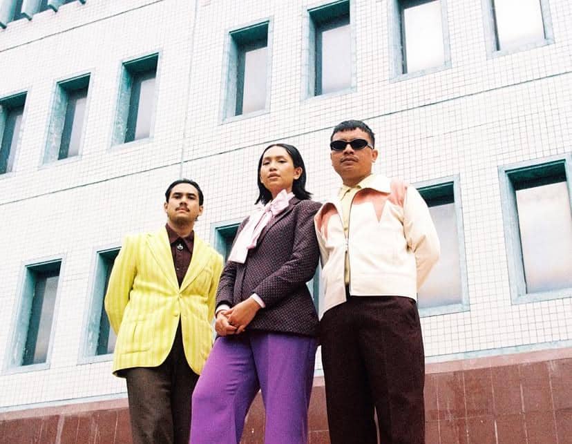 Tiga anggota Thee Marloes berpose di depan gedung berarsitektur modern dengan gaya busana retro berwarna cerah.