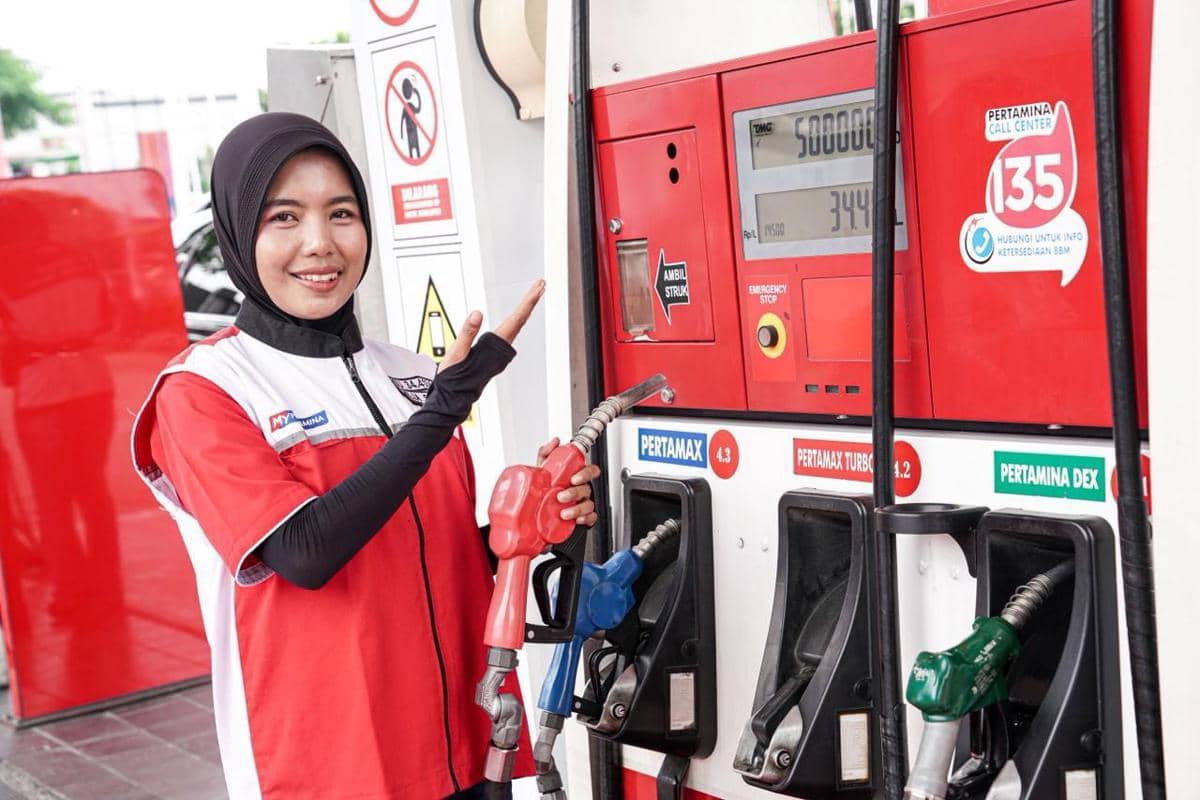 Petugas SPBU Pertamina mengenakan seragam merah putih sedang memegang nozzle bahan bakar dan menunjuk ke mesin pengisian BBM.
