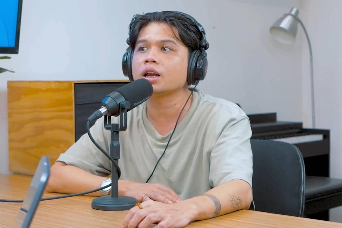 Uan Juicy Luicy mengenakan headphone dan berbicara di depan mikrofon di studio podcast dengan meja kayu dan lampu di latar belakang.