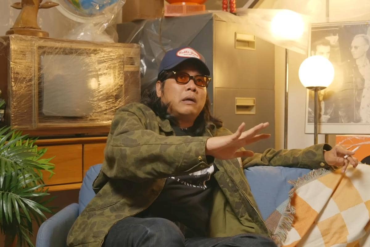 Gofar Hilman mengenakan jaket hijau bermotif loreng, topi biru, dan kacamata berwarna cokelat duduk di sofa biru di ruangan bergaya retro.