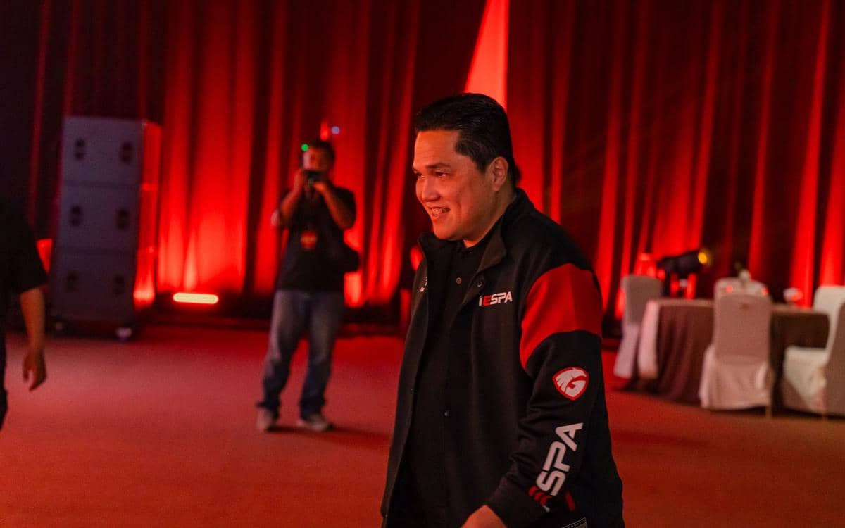 Seorang pria mengenakan jaket hitam merah bertuliskan IESPA berjalan di ruangan dengan pencahayaan merah dan latar tirai.