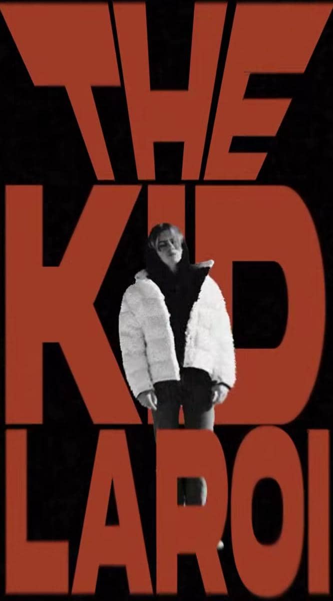 Seseorang mengenakan jaket tebal berwarna terang berdiri di depan latar hitam dengan tulisan besar berwarna merah bertuliskan THE KID LAROI.