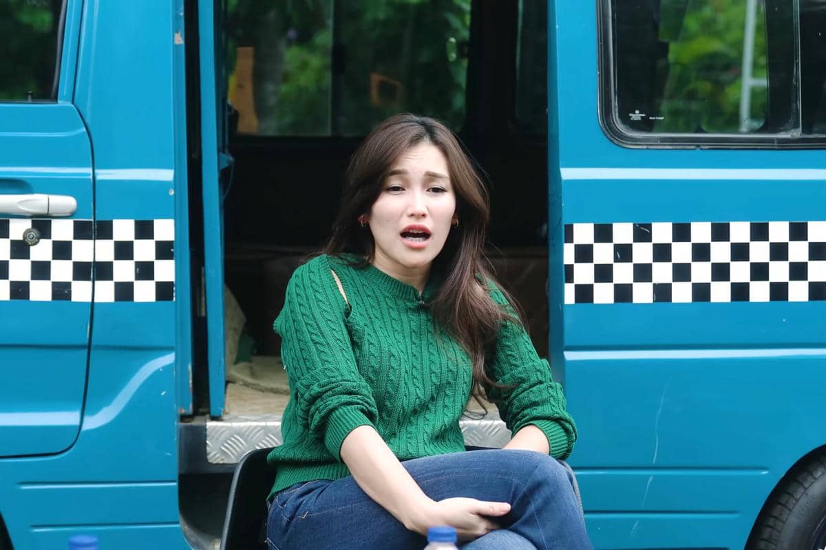 Ayu Ting Ting mengenakan sweter hijau duduk di depan kendaraan biru dengan pola kotak hitam putih di pintunya.