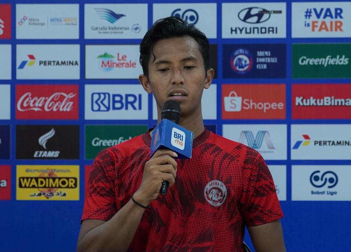 Seorang pemain Arema FC mengenakan jersey merah sedang berbicara memegang mikrofon di depan latar sponsor klub sepak bola.