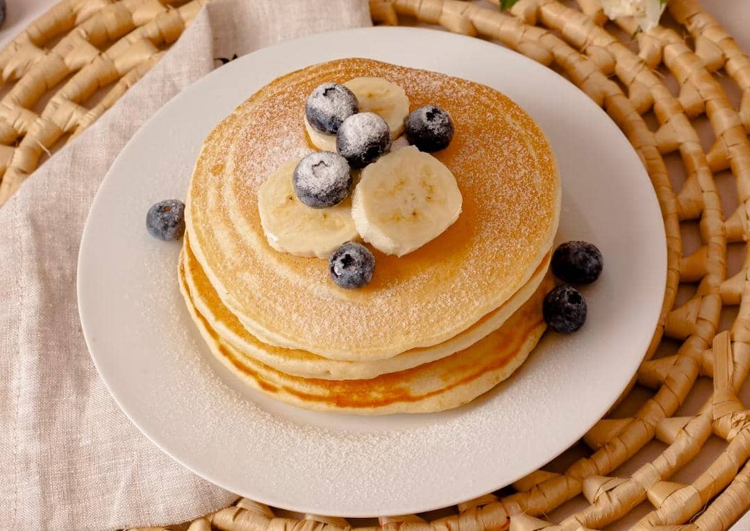 Tumpukan pancake lembut di atas piring putih dengan taburan gula halus, potongan pisang, dan buah blueberry segar.