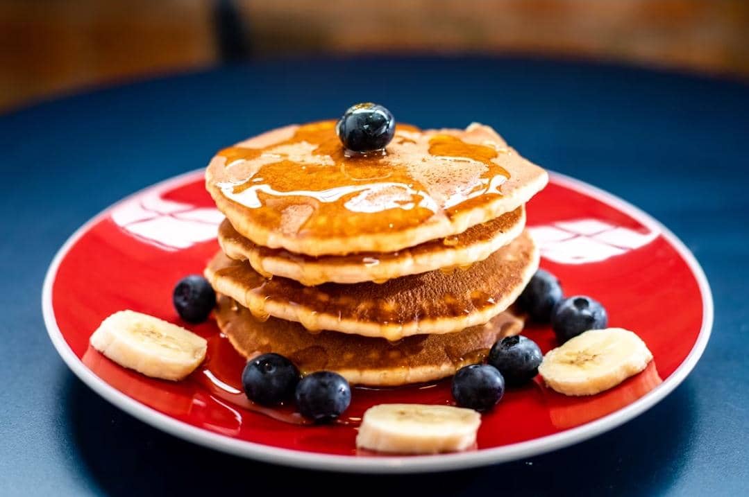 Tumpukan pancake dengan sirup madu di atas piring merah, dihiasi potongan pisang dan buah blueberry segar di sekelilingnya.