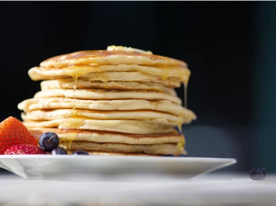 Tumpukan pancake lembut dengan sirup madu yang menetes, disajikan di atas piring putih bersama stroberi dan blueberry segar.