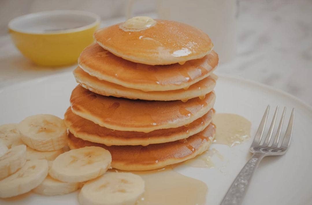 Tumpukan pancake lembut dengan mentega dan sirup madu di atas piring, disajikan bersama irisan pisang dan garpu di sampingnya.