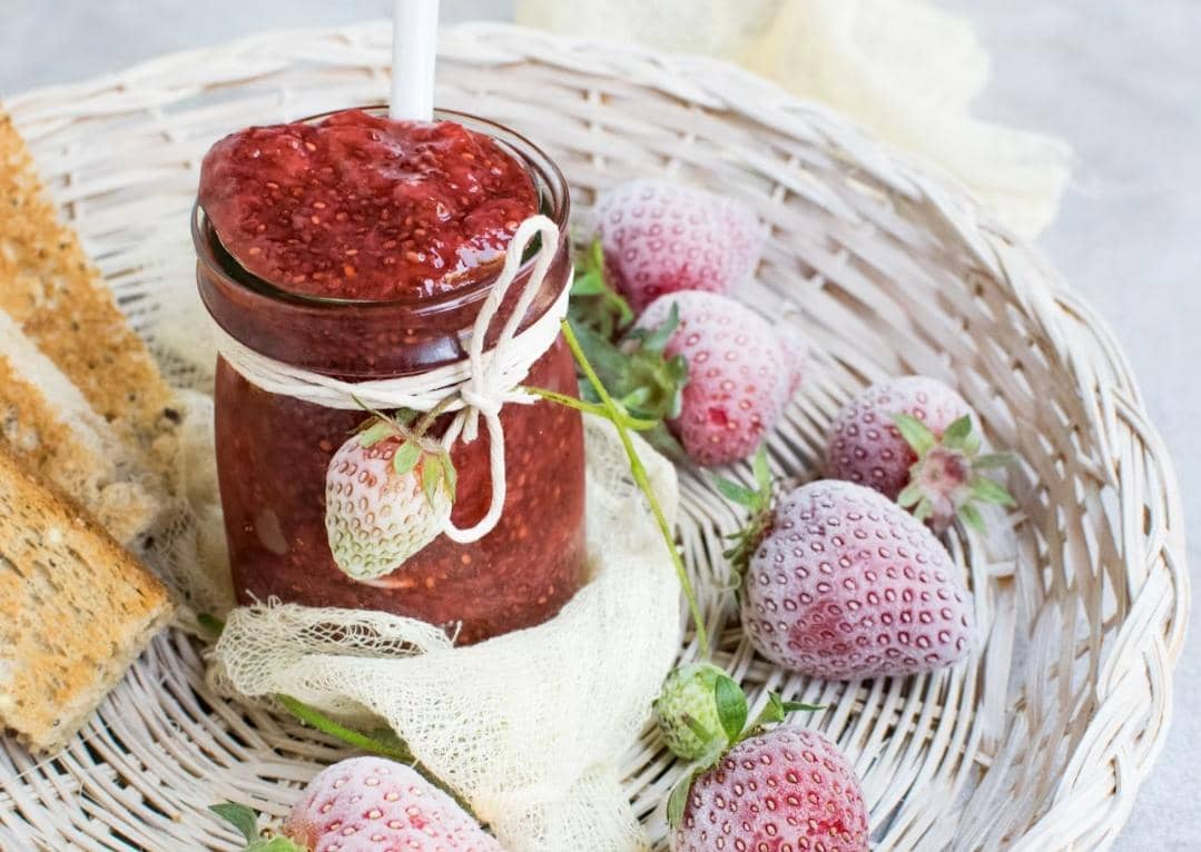 Stoples selai stroberi berwarna merah dengan buah stroberi segar sebagai ilustrasi makanan manis di meja.