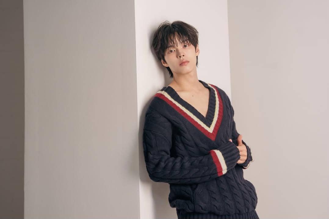 Seorang pria berpose bersandar di dinding dengan mengenakan sweater rajut biru tua bergaris merah dan putih di bagian leher.