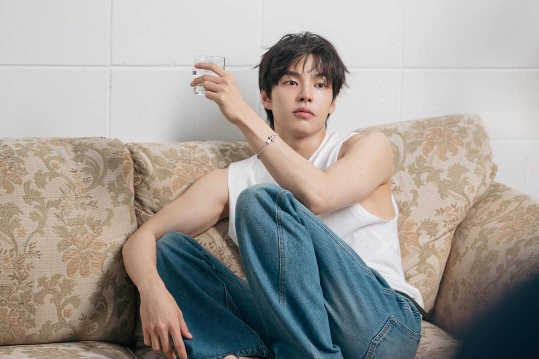 Seorang pria berambut hitam mengenakan kaus tanpa lengan putih dan celana jeans duduk santai di sofa sambil memegang gelas kecil.