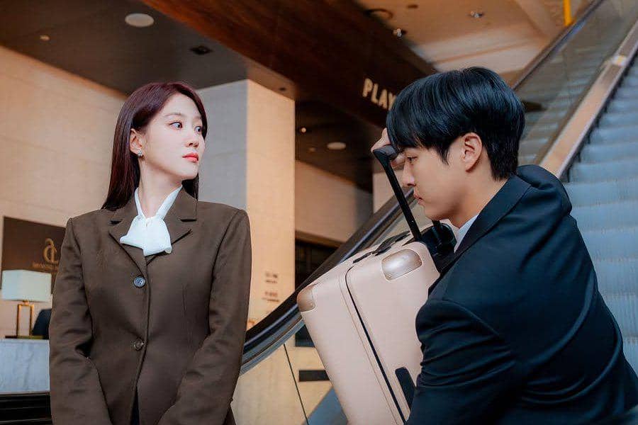 Dua pemeran drama Korea Spooky in Love berdiri di dekat eskalator, mengenakan setelan formal dengan koper berwarna krem.
