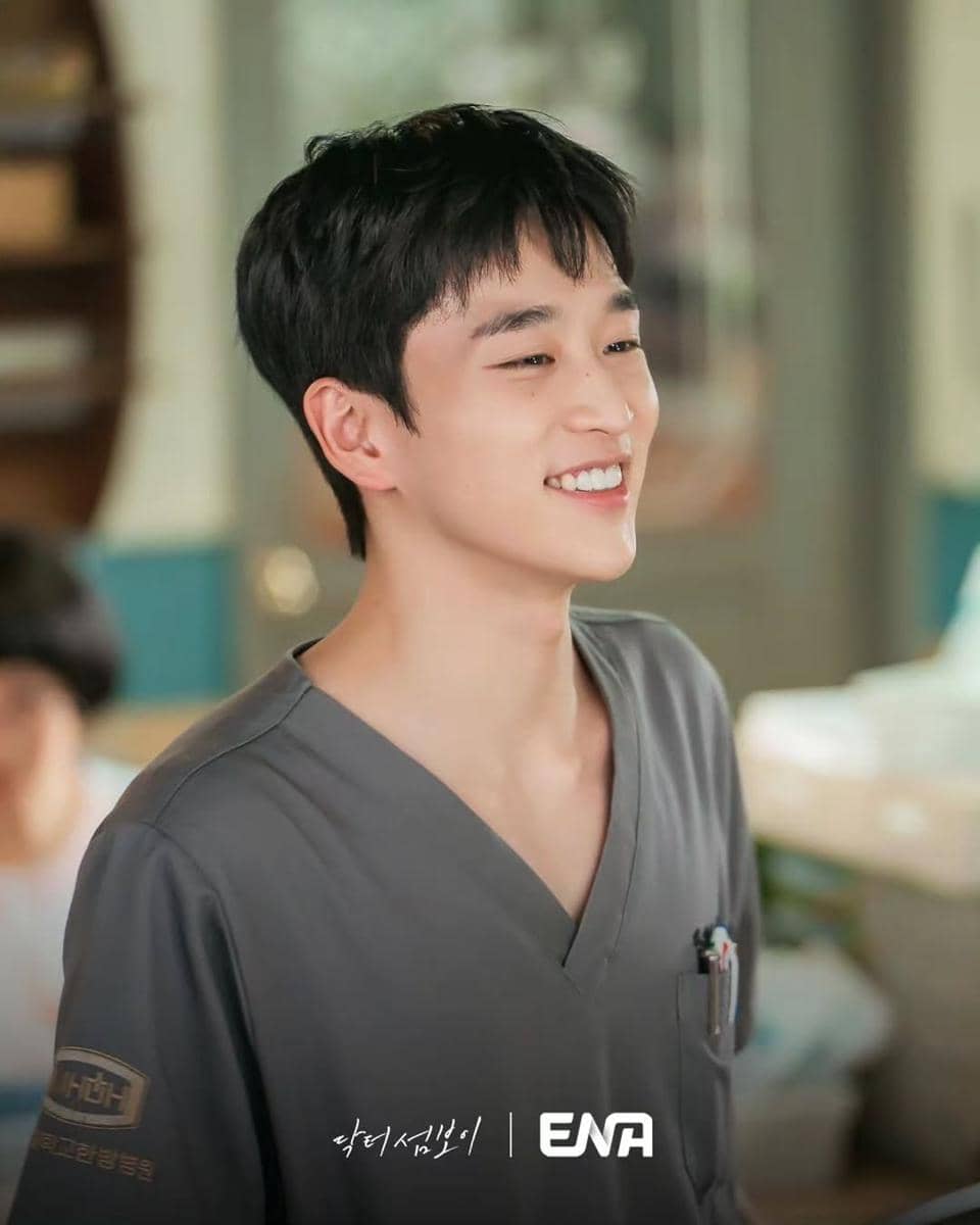 Seorang aktor mengenakan seragam medis abu-abu tersenyum cerah di lokasi syuting drama Doctor On The Edge.