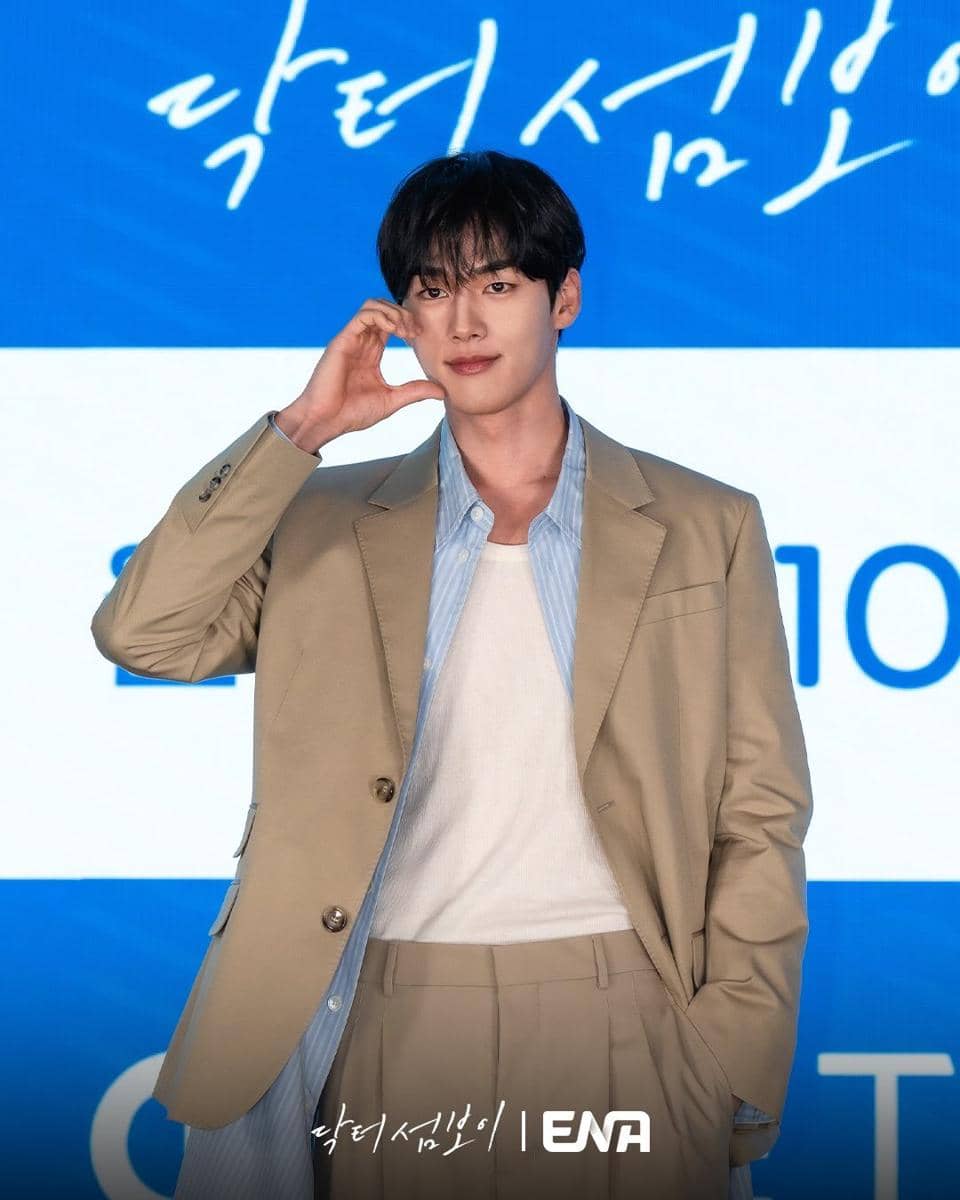 Seorang pria mengenakan setelan jas beige dengan kemeja biru muda dan kaus putih berpose di depan latar biru bertulisan ENA.