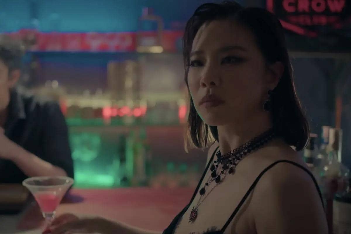 Seorang wanita dengan gaun hitam dan kalung perhiasan duduk di bar dengan pencahayaan neon dalam cuplikan drama The Crow Club.
