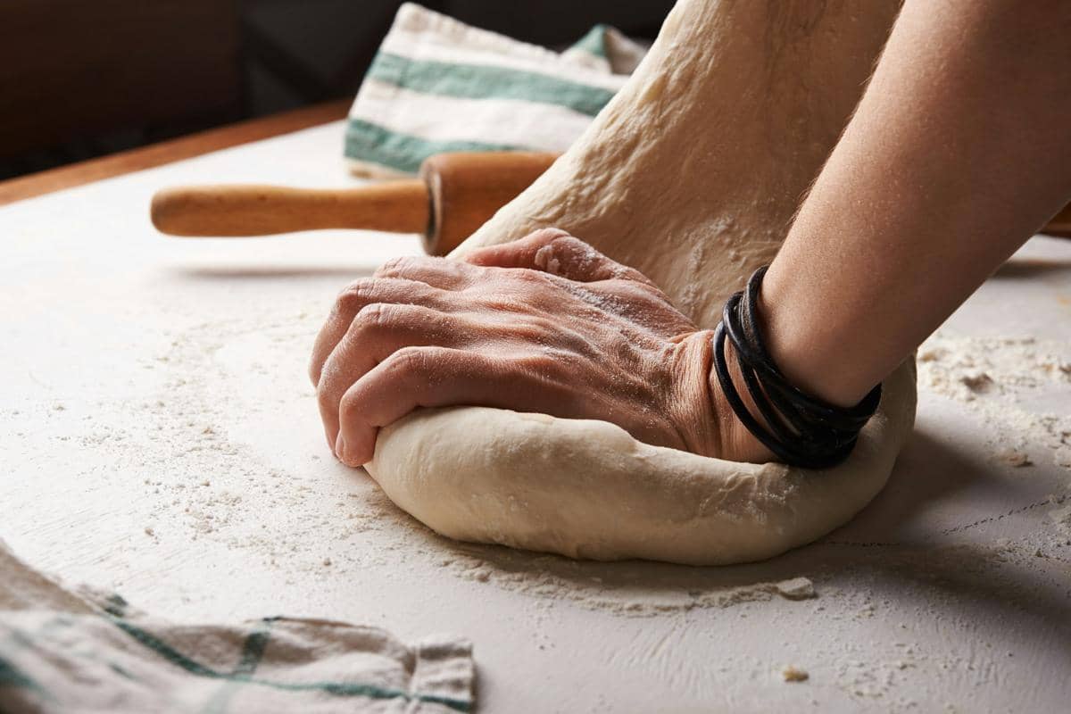 Tangan seseorang sedang menguleni adonan roti di atas meja dengan taburan tepung dan alat penggiling kayu di dekatnya.