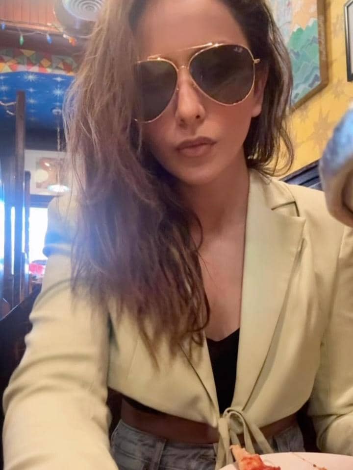 Seorang wanita mengenakan blazer krem dan kacamata hitam besar sedang duduk di restoran dengan latar warna-warni di dinding.