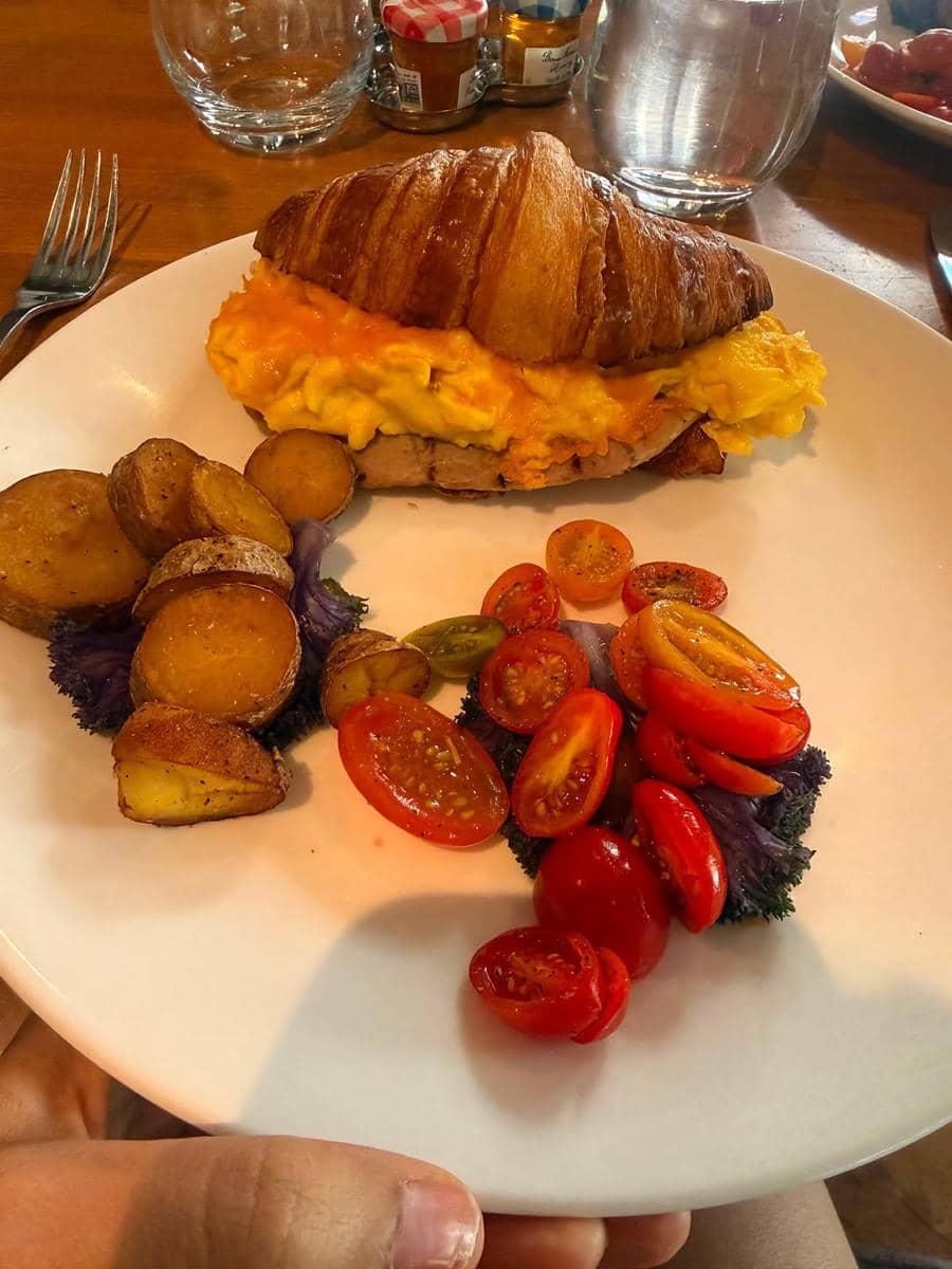 Sepiring croissant isi telur orak-arik disajikan dengan kentang panggang dan tomat ceri di atas meja kayu dengan gelas air di sampingnya.