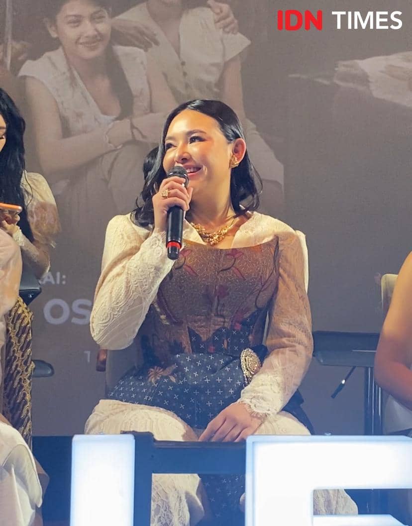 Seorang perempuan mengenakan gaun berwarna krem dan cokelat sedang tersenyum sambil memegang mikrofon di acara premier film di XXI Plaza Indonesia.