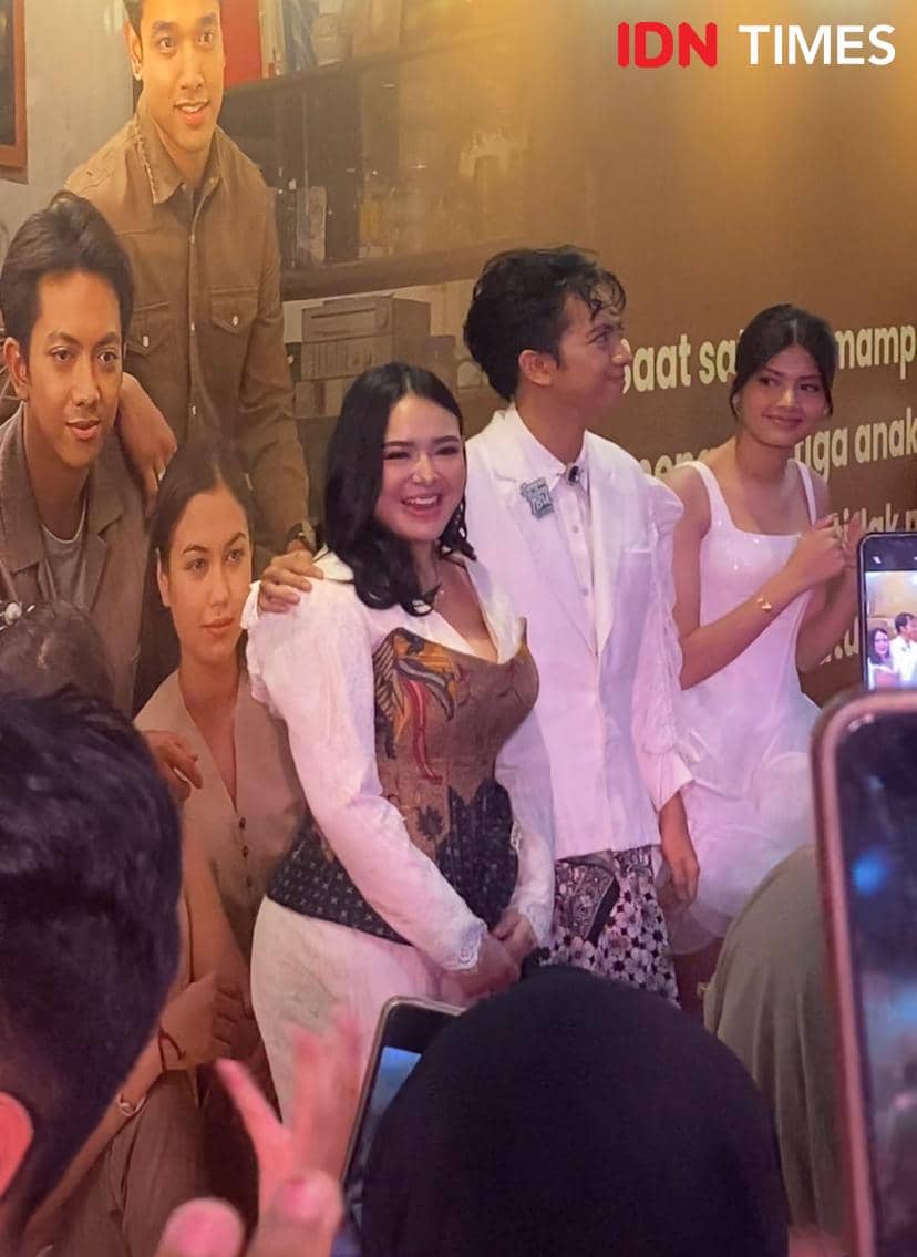 Seorang perempuan berbusana putih dengan korset bermotif batik berpose bersama dua orang muda di acara premier film di XXI Plaza Indonesia.