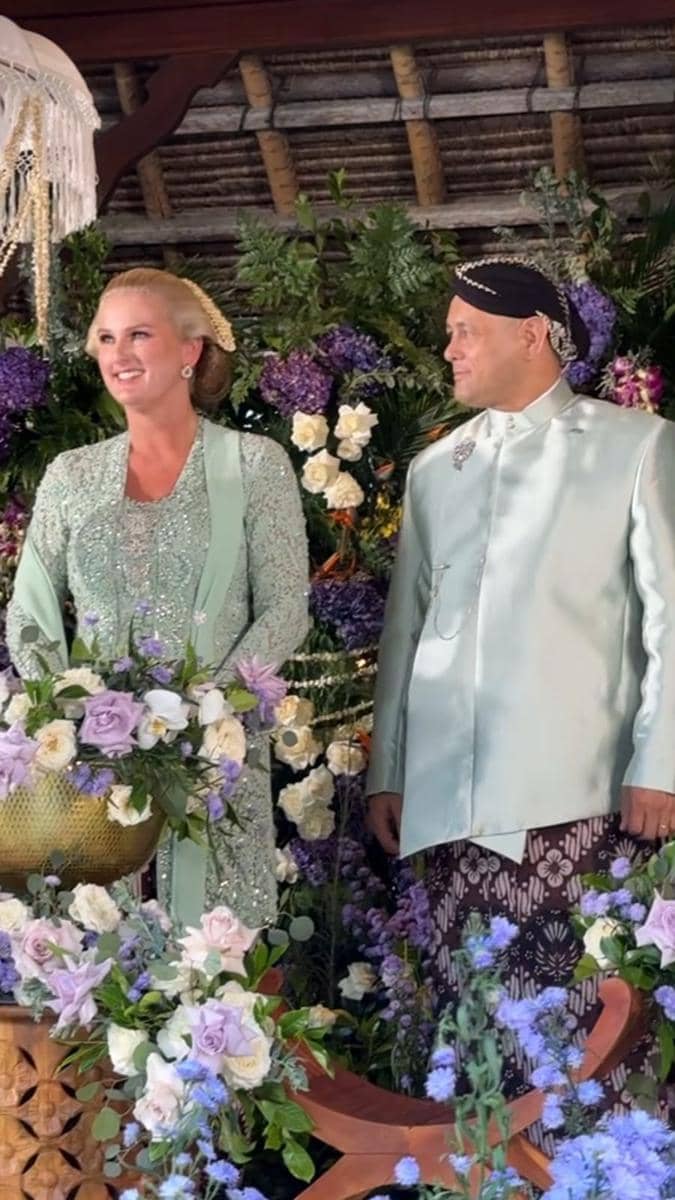 Sepasang tamu mengenakan busana tradisional berwarna hijau mint berdiri di depan dekorasi bunga ungu dan putih pada acara pengajian dan siraman.