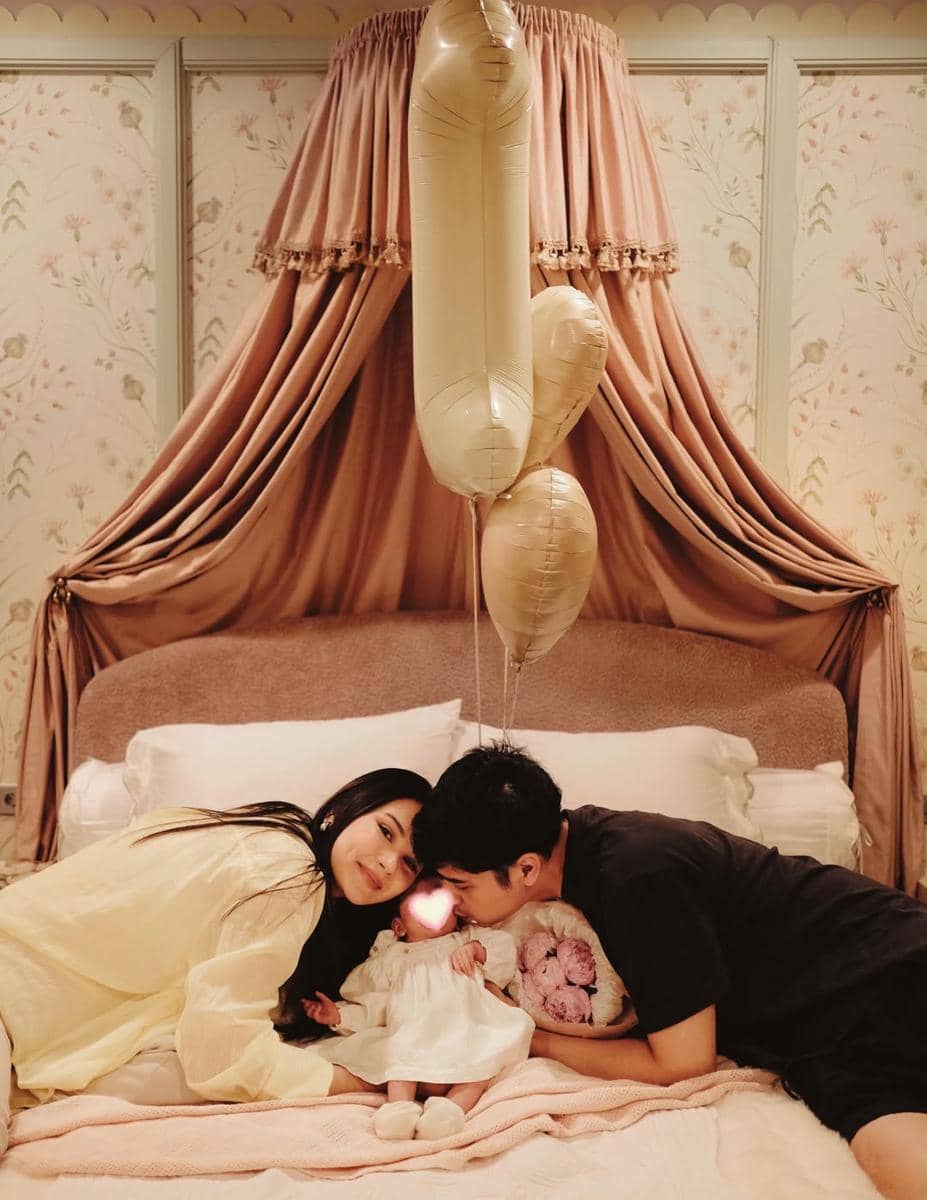 Pasangan berbaring di tempat tidur bersama bayi mereka yang mengenakan gaun putih, dikelilingi balon angka satu berwarna emas.