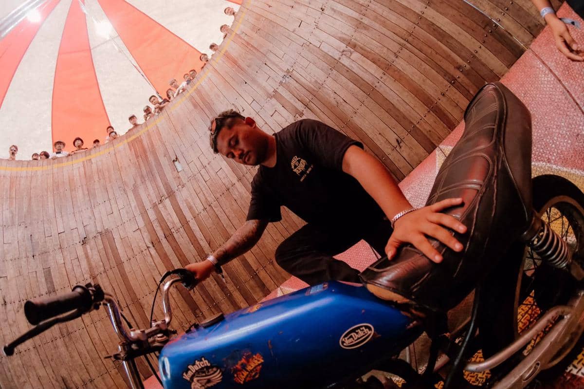 Seorang pria bersiap dengan motor biru di arena kayu berbentuk silinder dengan penonton mengelilingi di bagian atas tenda merah putih.