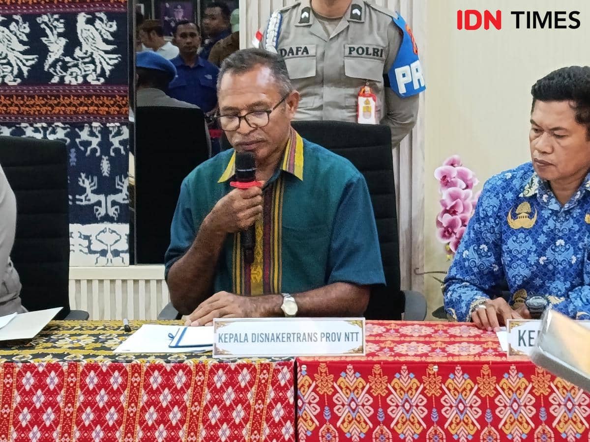 Seorang pejabat Disnakertrans NTT berbicara menggunakan mikrofon dalam rapat resmi bersama sejumlah peserta di ruangan berdekorasi kain tenun.