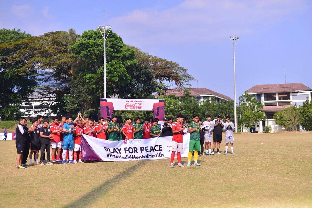 Para pemain sepak bola universitas berpose bersama di lapangan sambil memegang spanduk Play for Peace dalam Liga Universitas Coca Cola 2026.