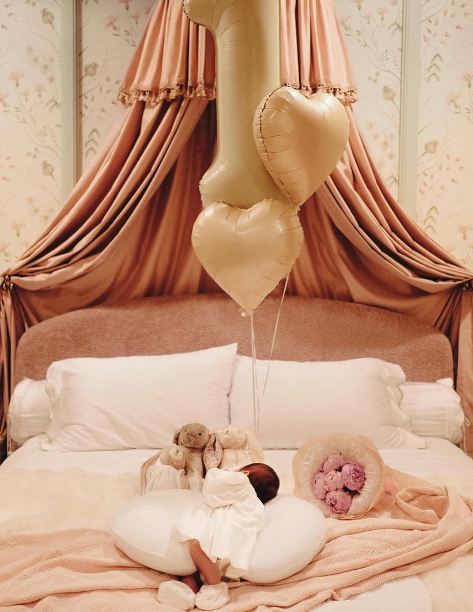 Bayi berbaring di atas tempat tidur dengan balon angka satu dan dua balon hati berwarna emas serta buket bunga di sampingnya.