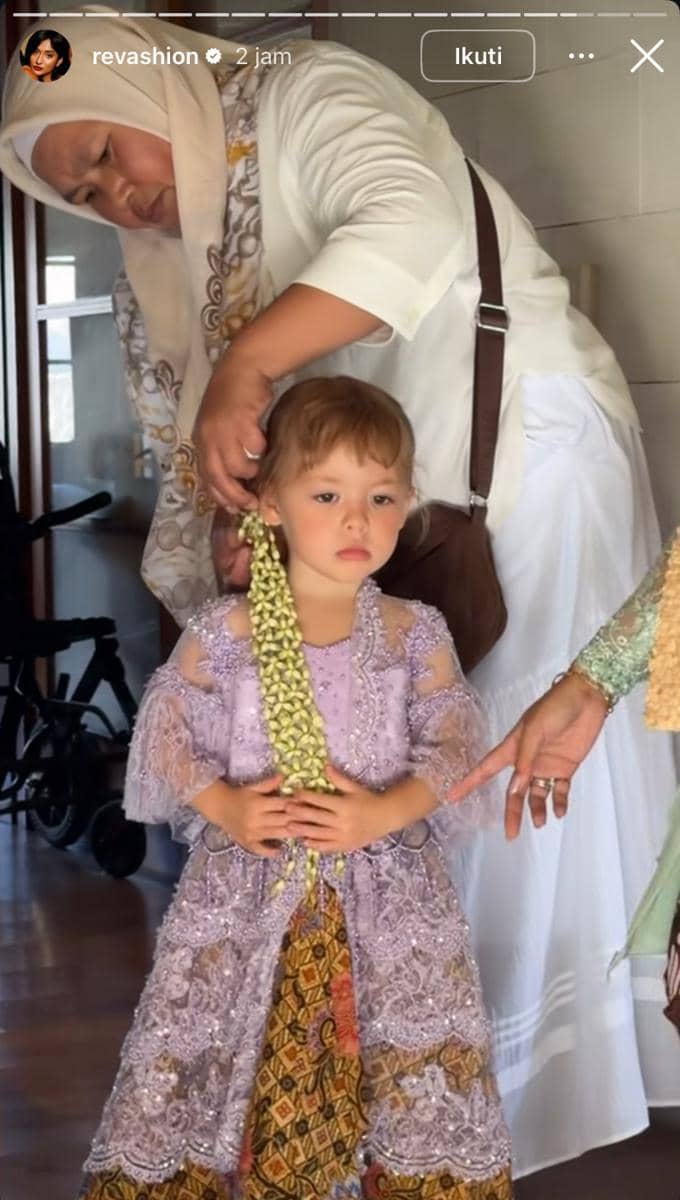 Seorang anak perempuan mengenakan kebaya ungu dan kain batik sedang dipasangkan hiasan bunga melati di rambutnya oleh seorang wanita berhijab.