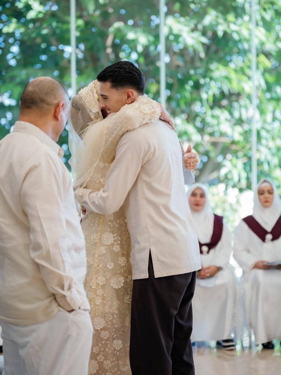 Seorang pria memeluk wanita berhijab dalam balutan kebaya krem di acara pengajian dan siraman dengan latar pepohonan hijau.
