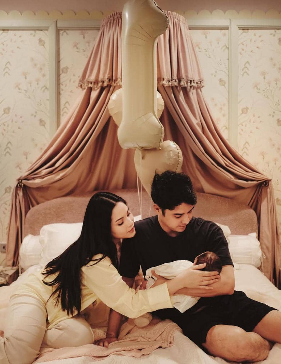 Pasangan muda merayakan satu bulan kelahiran bayi mereka di kamar dengan dekorasi balon angka satu berwarna emas di atas tempat tidur.