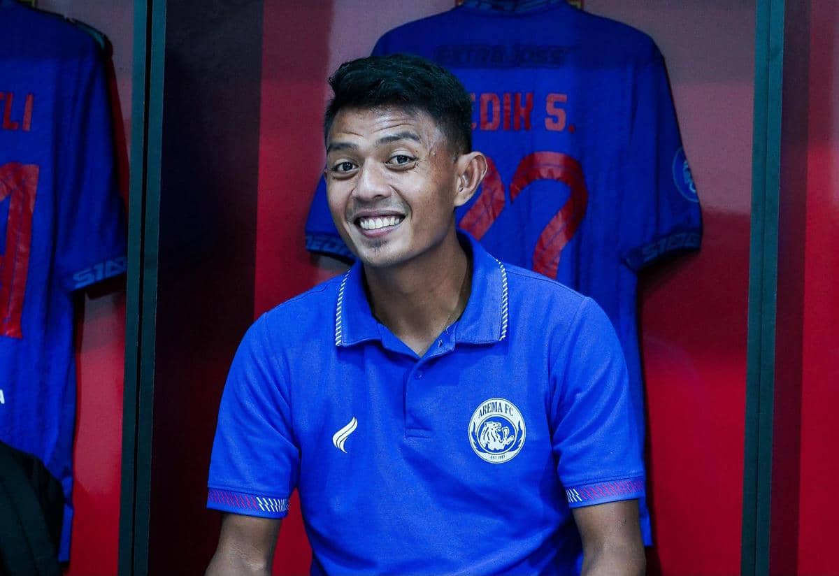 Seorang pemain Arema FC mengenakan seragam biru dengan logo klub, tersenyum di ruang ganti dengan latar belakang jersey tim.