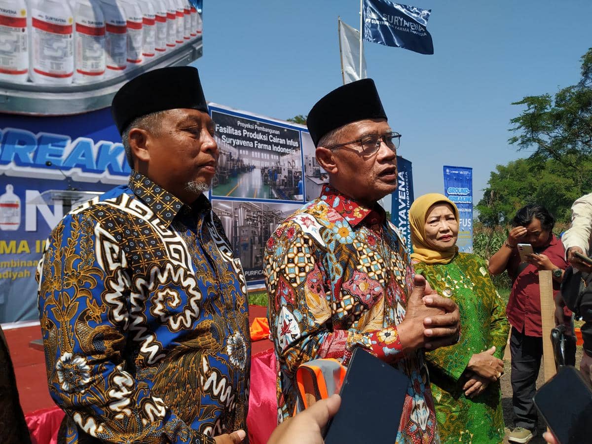 Seorang pria berbaju batik berbicara kepada media di acara luar ruangan bersama dua orang lainnya dengan latar spanduk dan bendera perusahaan.