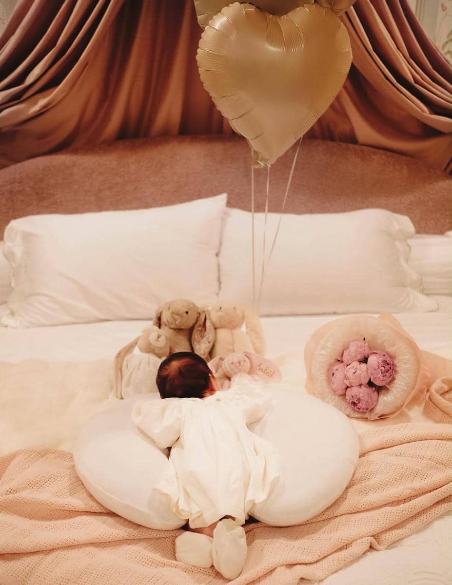 Bayi mengenakan pakaian putih berbaring di atas bantal berbentuk donat dengan balon hati emas dan boneka di tempat tidur bernuansa pastel.