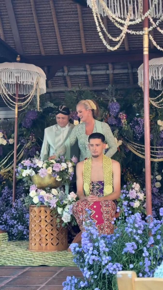 Seorang pria mengenakan busana adat Jawa duduk di depan pasangan yang mengenakan pakaian tradisional hijau muda dalam acara siraman dengan dekorasi bunga ungu dan putih.