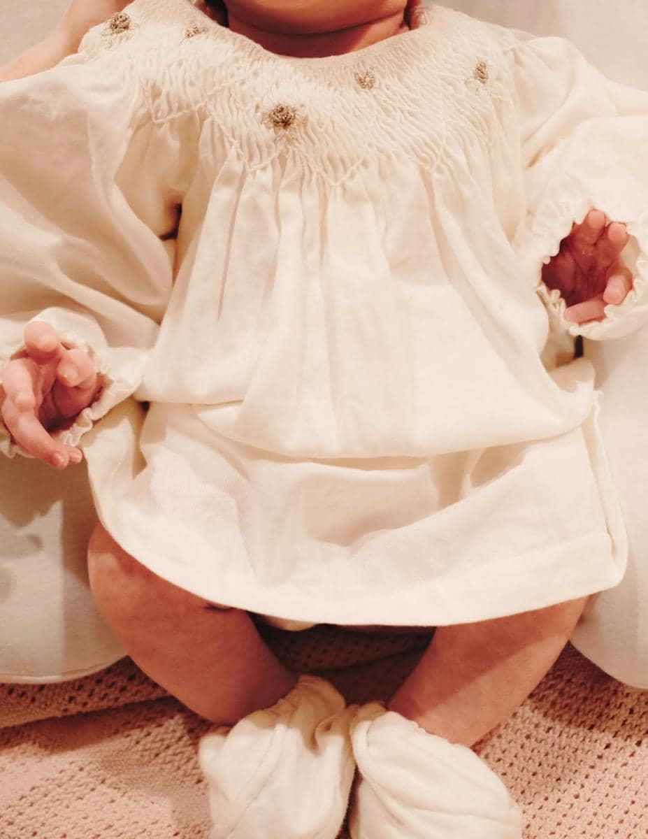 Bayi mengenakan gaun putih lembut dengan detail bordir halus dan sepatu kain, duduk di atas permukaan kain berwarna pastel.