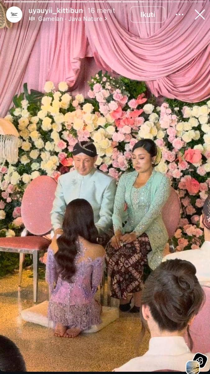 Seorang perempuan mengenakan kebaya ungu berlutut di hadapan dua orang yang duduk di kursi, dengan latar bunga dan tirai merah muda.