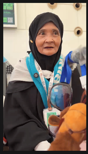 Seorang jamaah haji perempuan mengenakan jilbab hitam dan syal biru muda di Bandara King Abdulaziz, Jeddah, bersiap pulang ke Sulawesi Selatan.