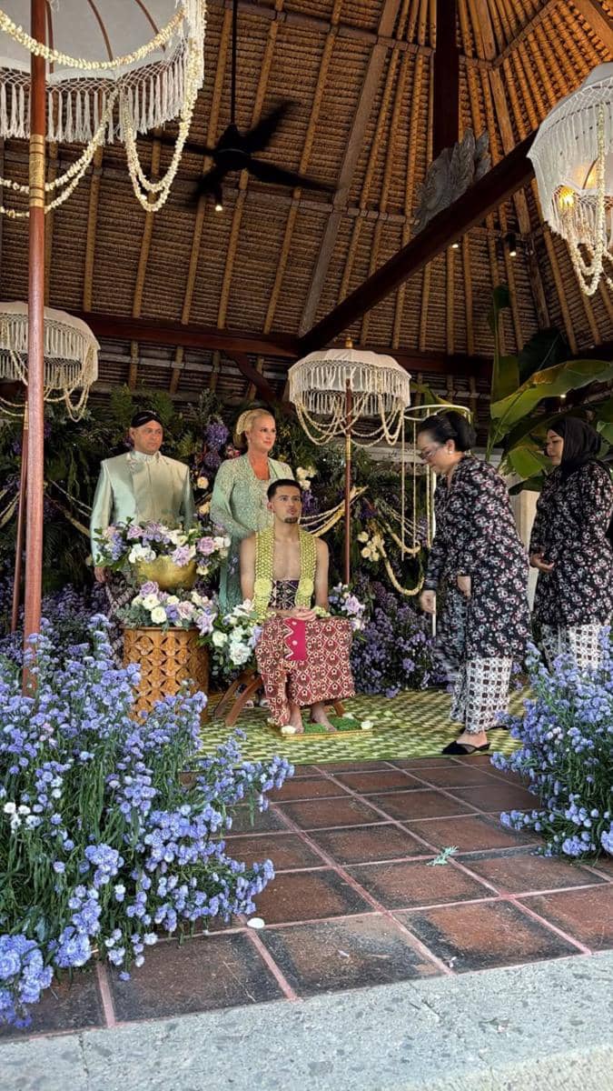 Suasana acara pengajian dan siraman dengan dekorasi bunga ungu, seorang pria duduk mengenakan kain batik dan selendang kuning di pelaminan tradisional.