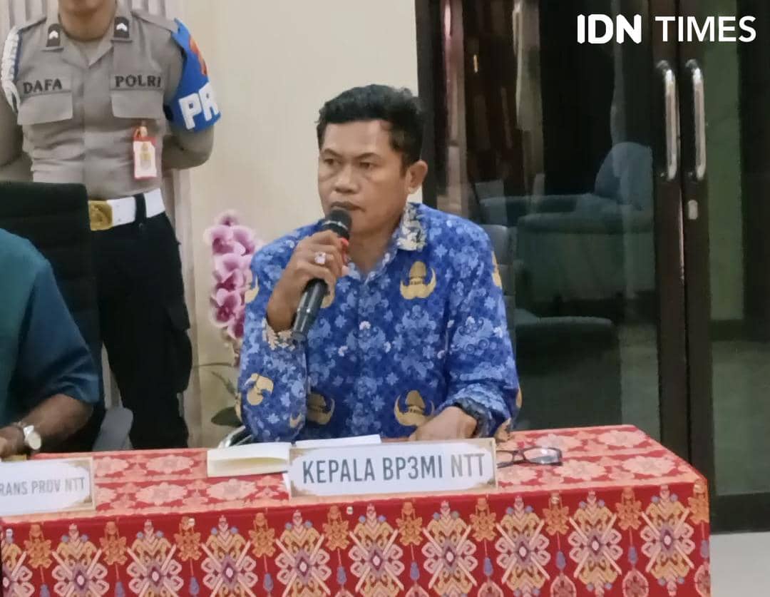 Seorang perwakilan BP3MI NTT mengenakan batik biru berbicara menggunakan mikrofon di meja rapat dengan papan nama jabatannya di depan.