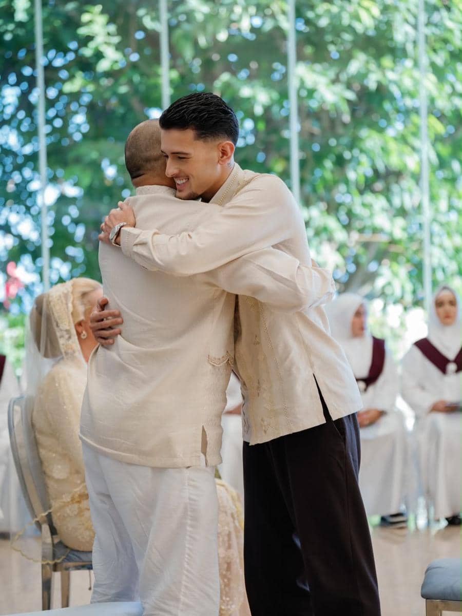 Seorang pria mengenakan baju tradisional berwarna krem memeluk pria lain dalam acara pengajian dan siraman keluarga di ruangan bercahaya alami.