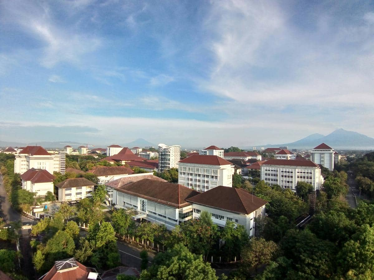 Pemandangan udara kompleks Universitas Gadjah Mada di Sleman dengan deretan gedung beratap merah dan latar pegunungan di bawah langit cerah.