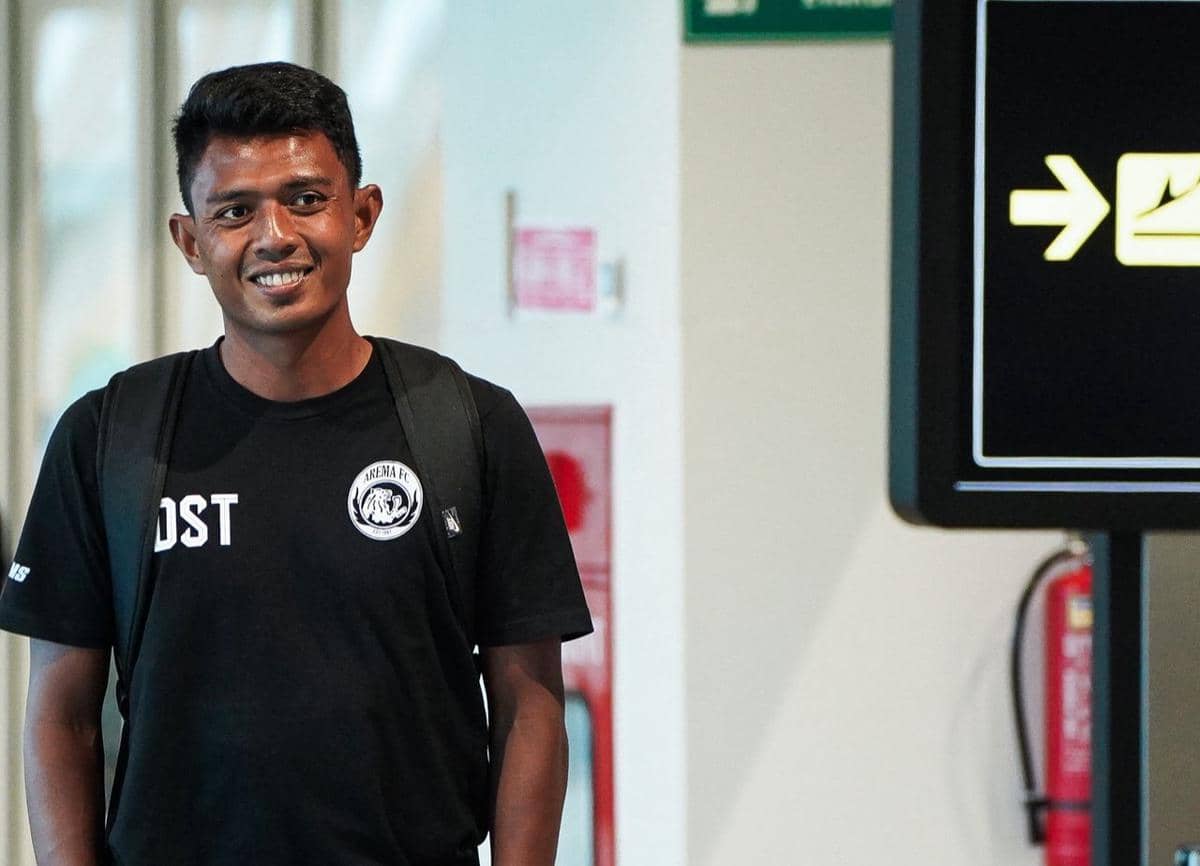 Seorang pemain Arema FC mengenakan kaus hitam bertuliskan logo klub dan membawa ransel hitam di area dalam gedung.