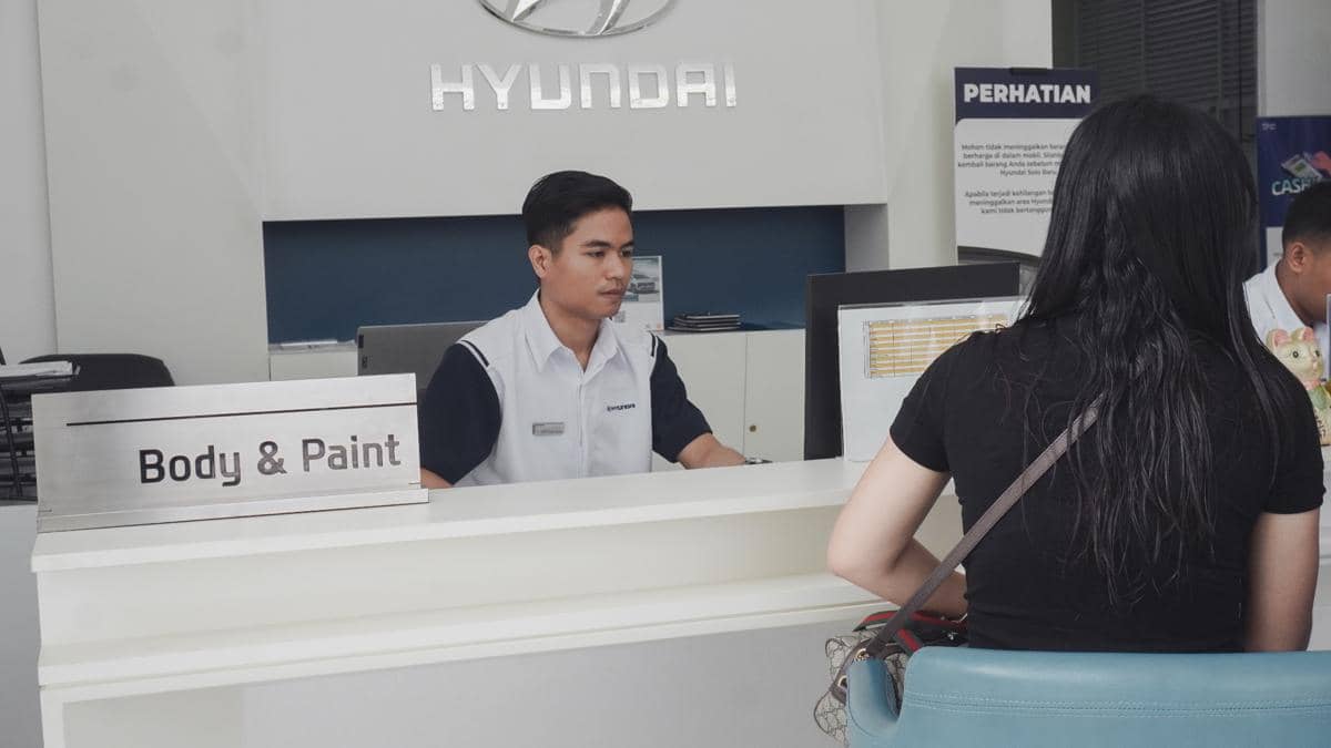 Petugas layanan body and paint Hyundai melayani pelanggan di meja resepsionis dengan suasana profesional di ruang pelayanan.