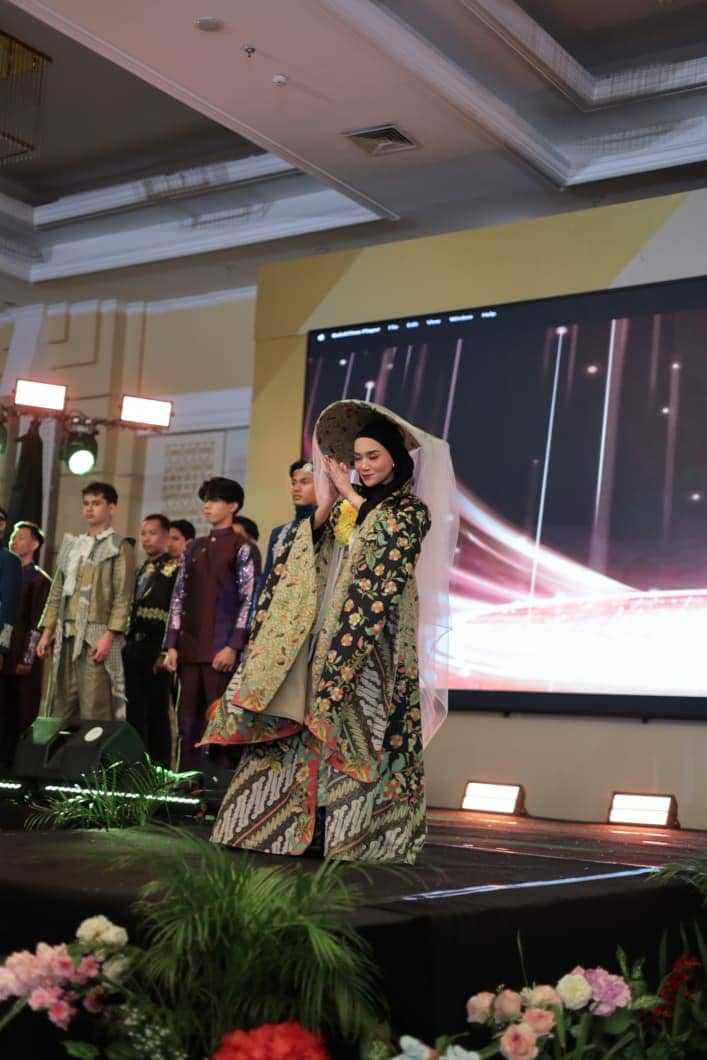 Seorang perempuan mengenakan busana tradisional berhijab dengan motif batik dan songket berjalan di atas panggung dalam acara resmi di dalam ruangan.