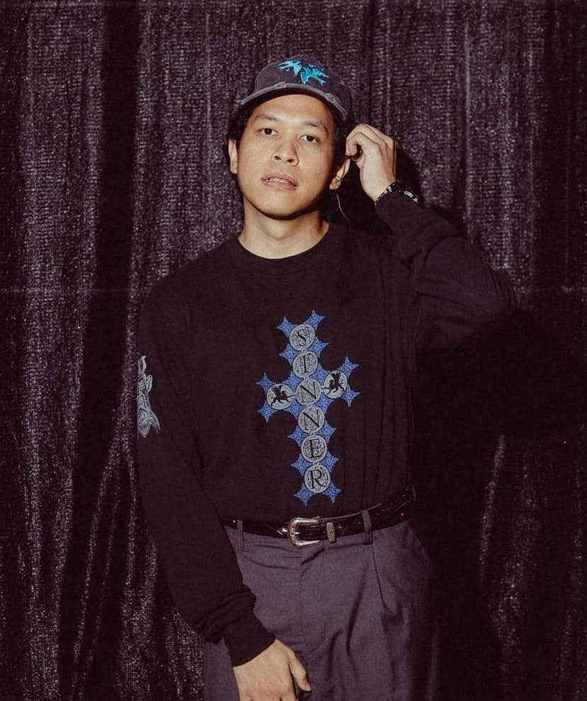 Seorang pria mengenakan topi abu-abu dan kaus hitam bergambar salib biru dengan tulisan, berdiri di depan tirai hitam.