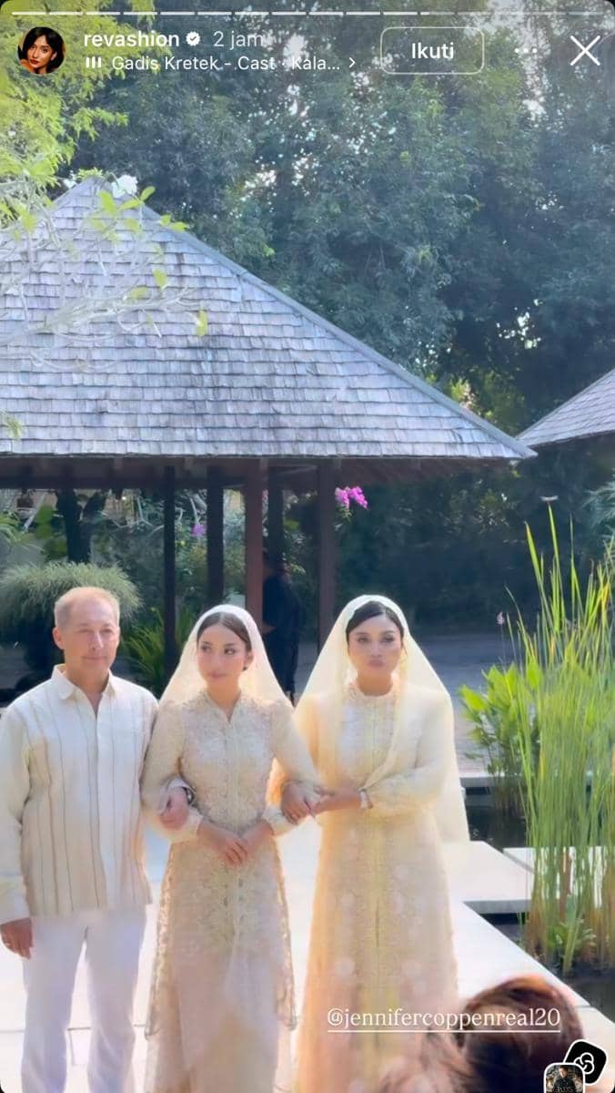 Seorang perempuan mengenakan kebaya krem bersama dua orang lainnya berjalan di area taman dengan latar bangunan beratap kayu.